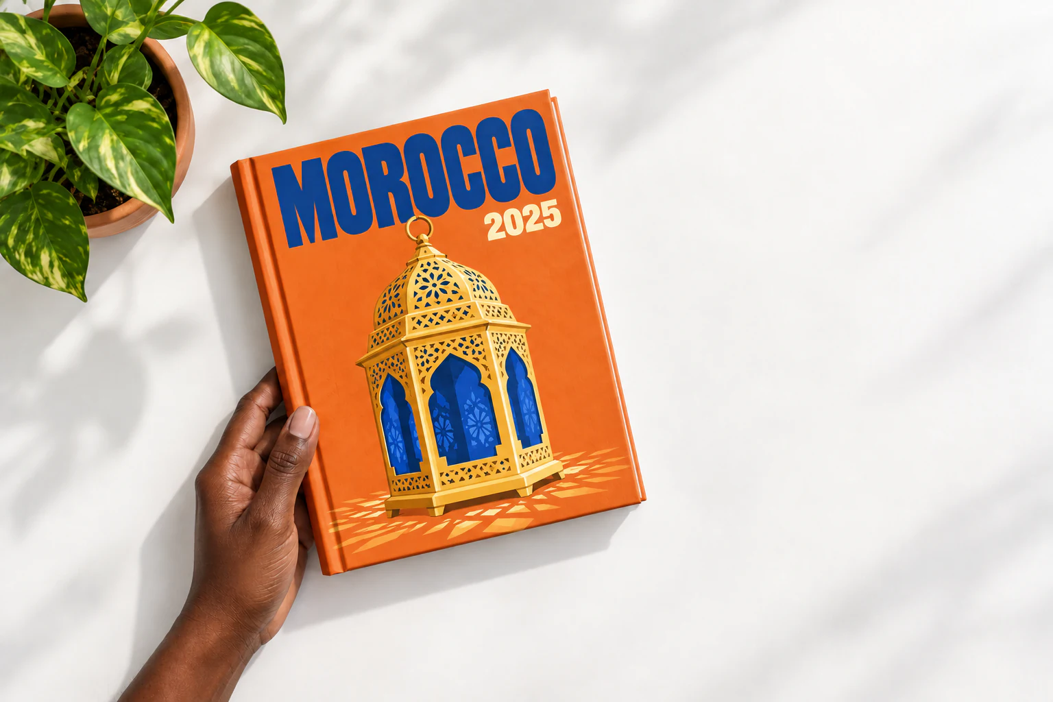 Overhead view of a Black woman's hand holding the Morocco photo album above a white table with a potted plant